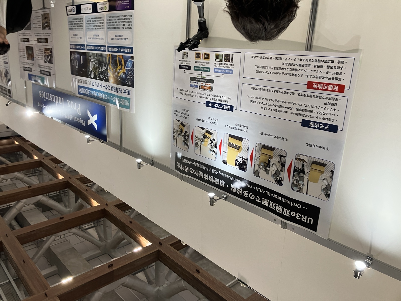 Prox Industriesの展示パネル。UR3e双腕ロボットによる箱詰め工程と、CV・Motion Planning・VLA・RLを組み合わせる構成を説明