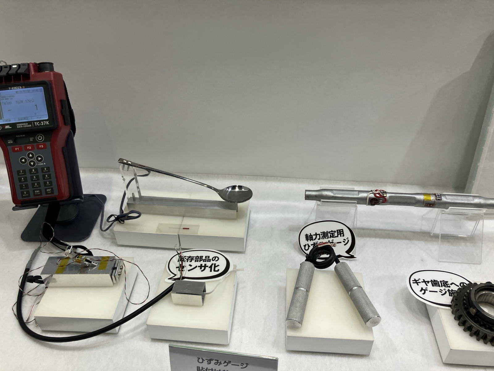 東京測器研究所のひずみゲージ展示。スプーン、既存部品、軸力測定用部材、ギヤへの貼付例